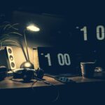 A dimly lit modern office desk with a laptop, headphones, and speakers at 1:00 AM.